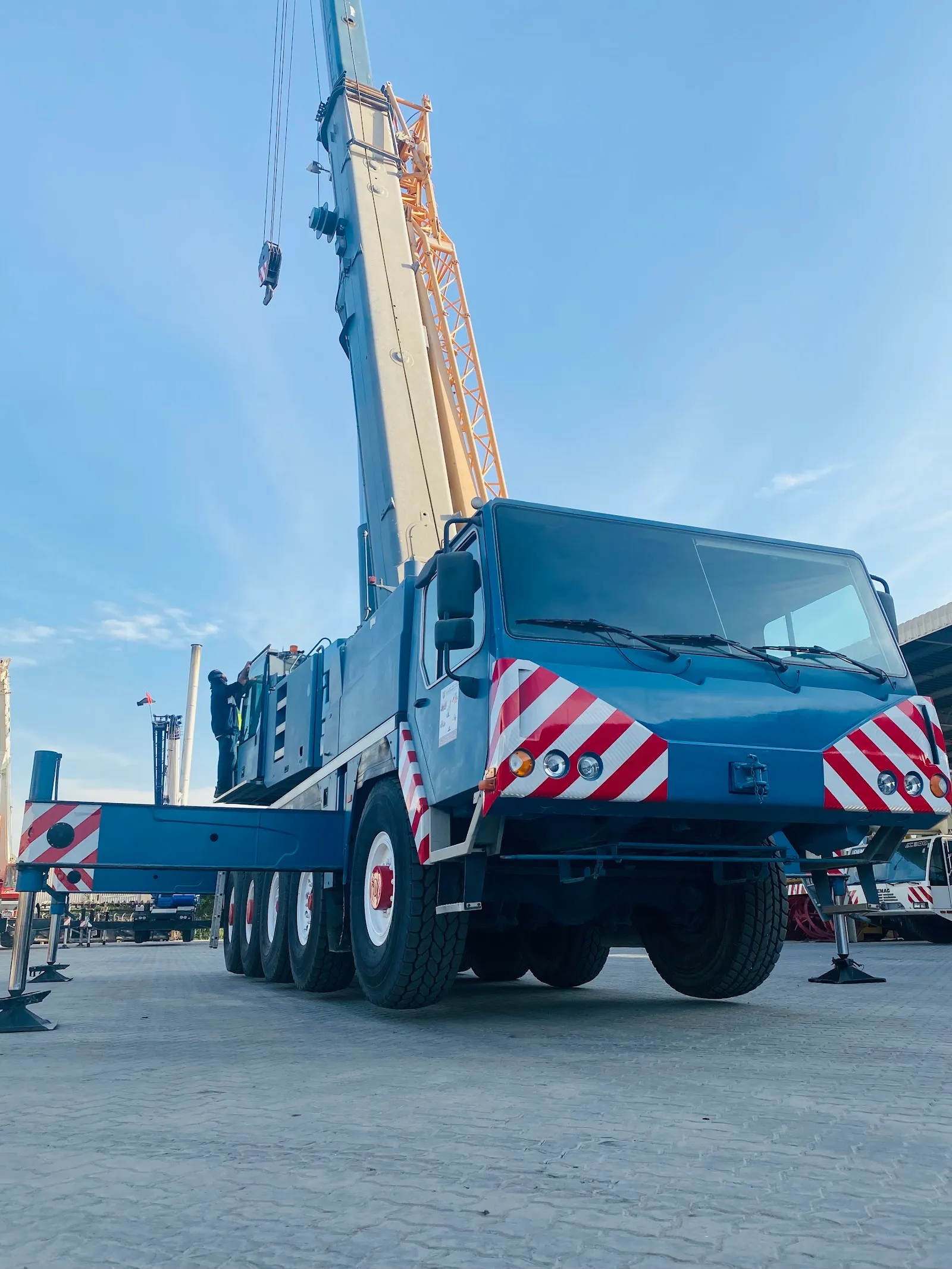 A blue five-axle all-terrain mobile crane on outriggers with a lattice jib extension fitted, an operator climbing to the cab, in a port yard.
