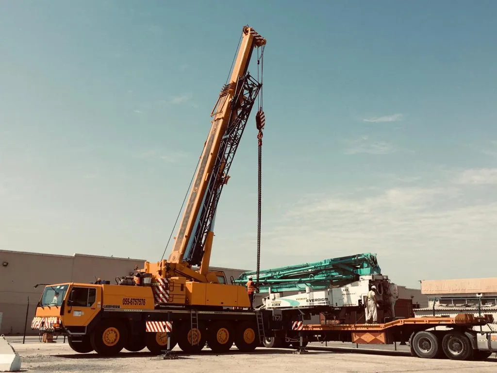 A Liebherr all-terrain mobile crane lifting a concrete pump boom onto a lowbed trailer in a port yard, with an operator standing on the trailer deck.