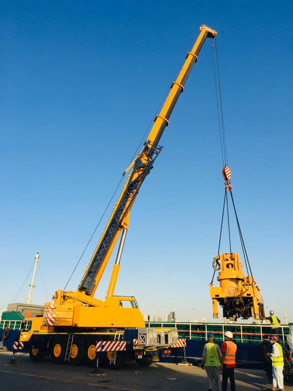 A Liebherr mobile crane with its boom fully extended lifting a heavy machine on chains off a vessel at the quay, with riggers in high-visibility vests watching from the deck.
